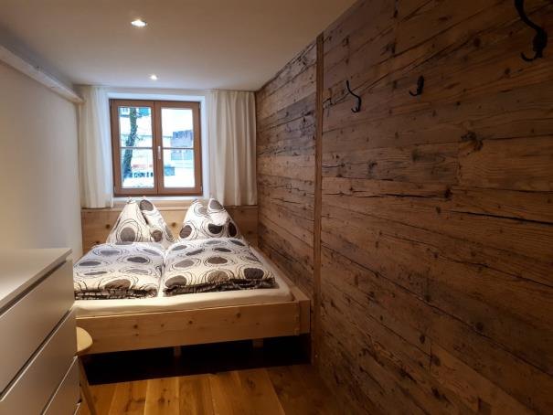 Doppelbett vor einer Wand aus altem Holz, daneben ein Fenster mit Vorhang